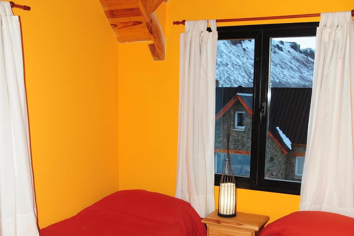 Quarto com duas camas, uma mesa de cabeceira de madeira, janela com cortinas e vista para uma casa.