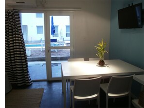 Un comedor con una mesa y sillas blancas, una planta en maceta y vistas a una zona de estar exterior a través de un ventanal.