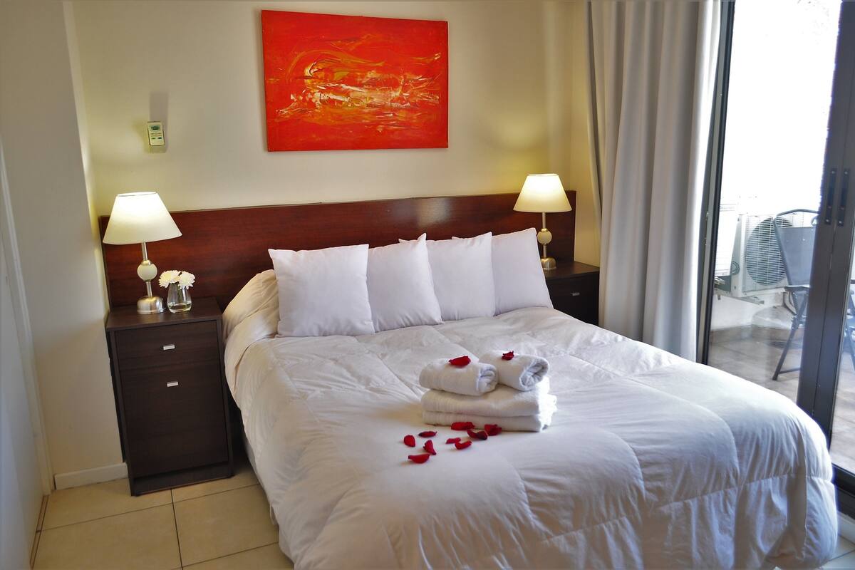 Habitación de hotel con una cama grande, dos lámparas de noche, un cuadro abstracto rojo y vistas al exterior.