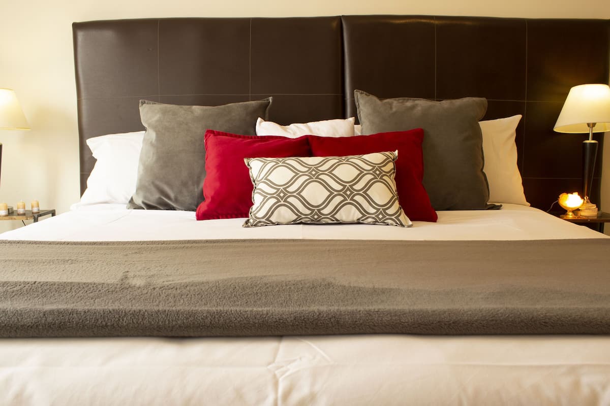 Una cama bien tendida con un cabecero oscuro, varios almohadones y una mesita de noche con una lámpara.