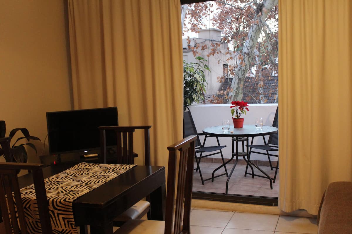 Una sala de estar con televisor, sofá, mesa de comedor con sillas y balcón con planta en maceta.