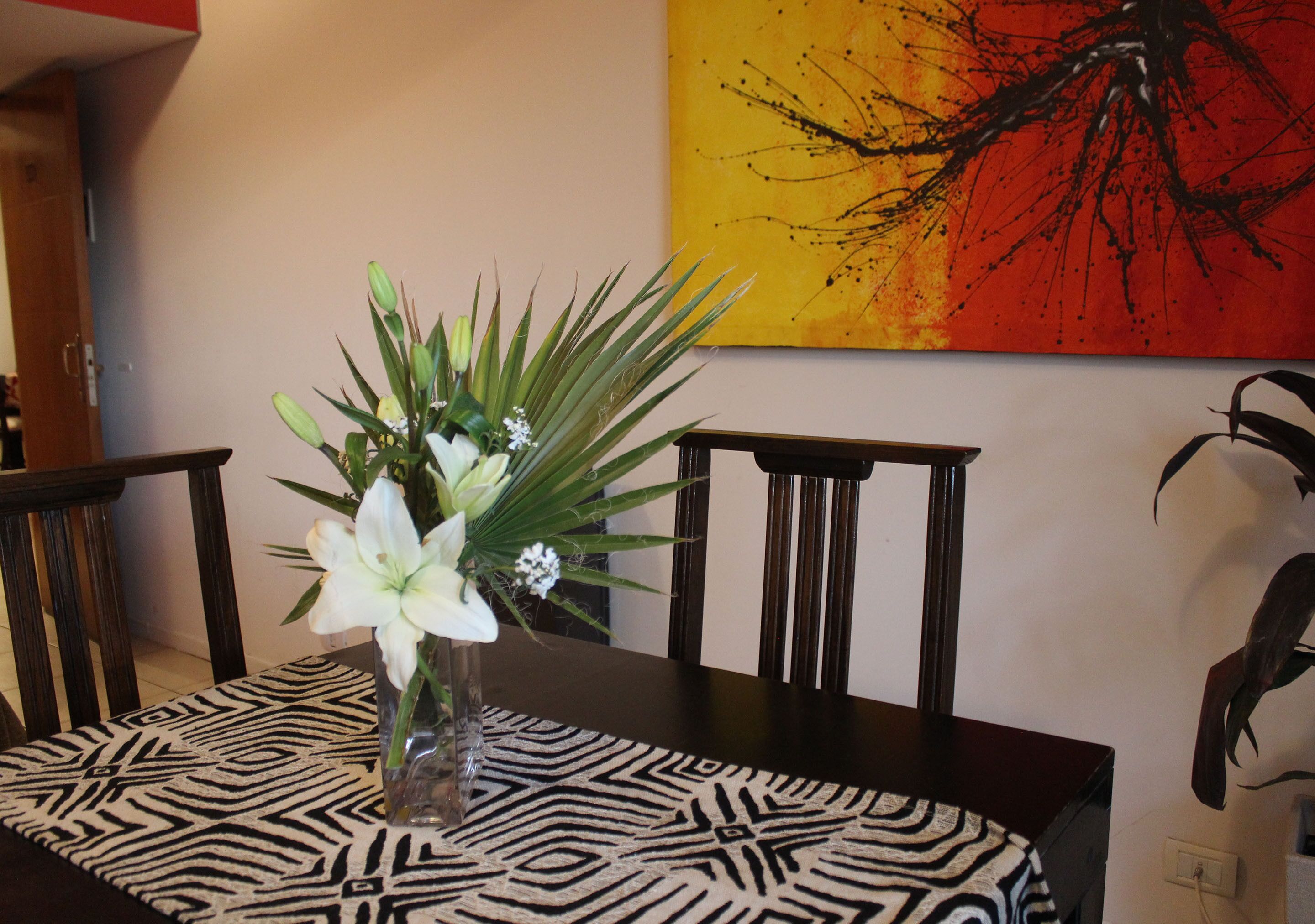 Une table à manger agrémentée d’un chemin de table à motifs, un vase avec des fleurs blanches et un tableau accroché au mur.