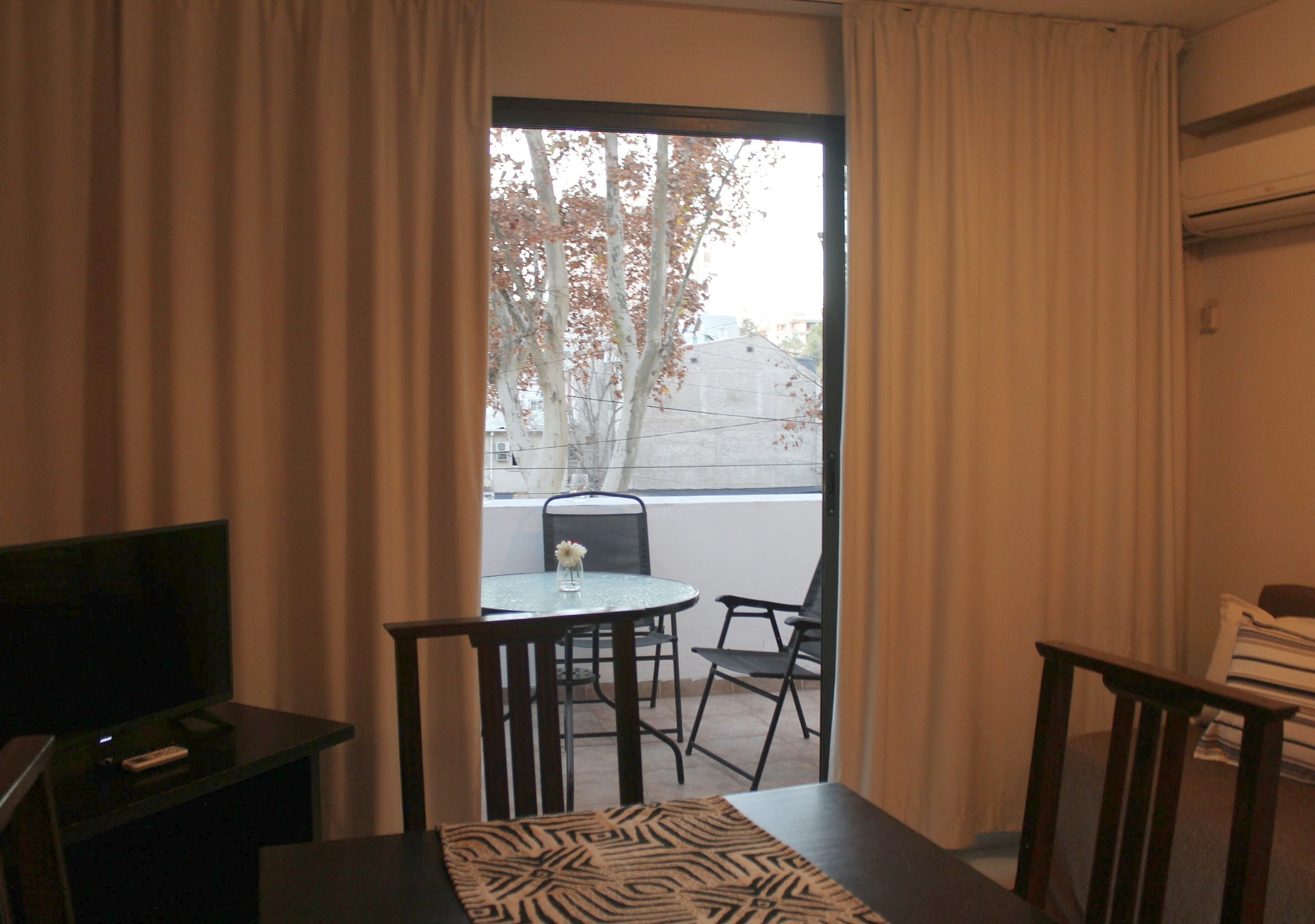 Een kamer met balkon, tafel, stoelen, televisie en gordijnen.