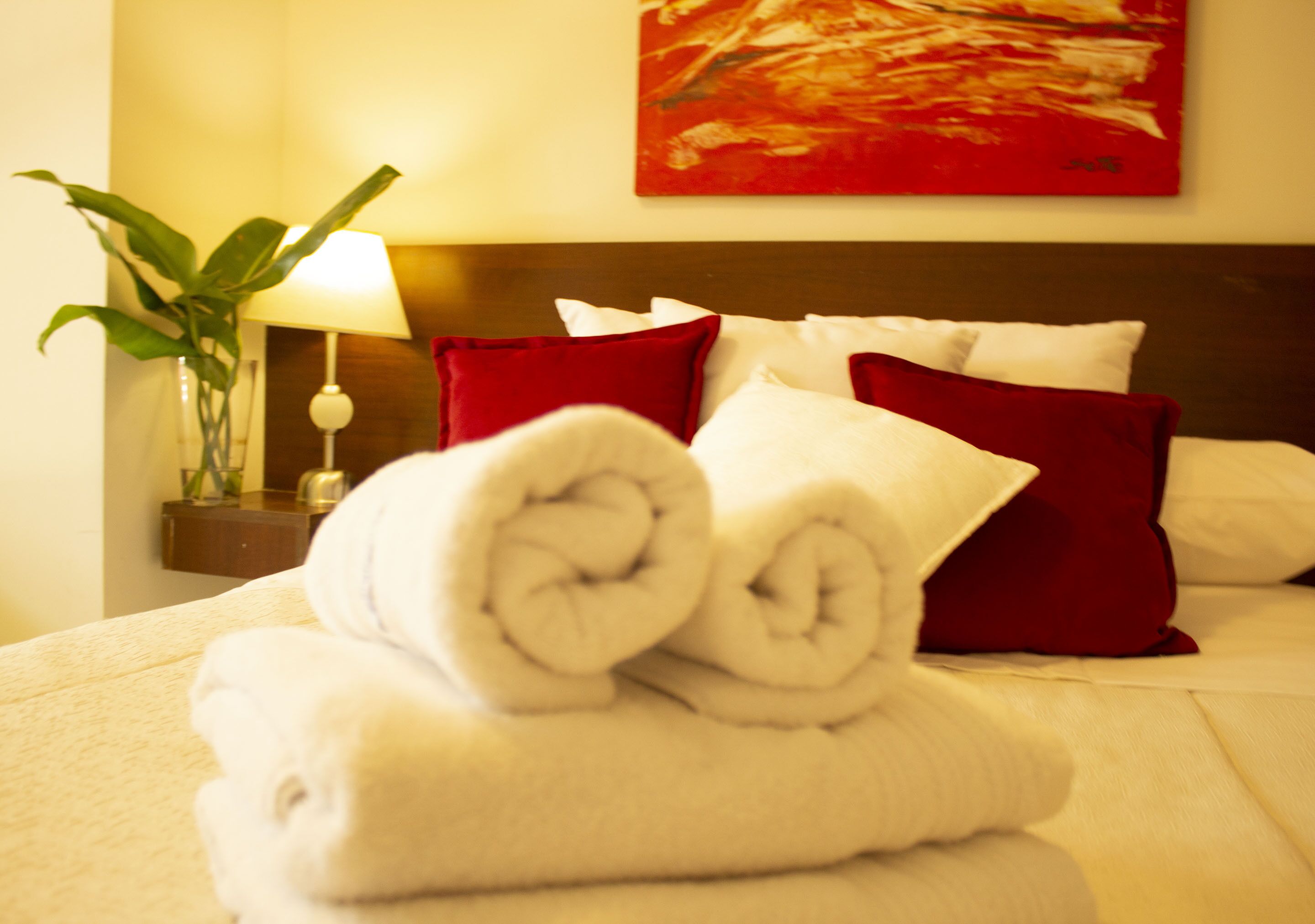 Una cama bien hecha con toallas blancas enrolladas y almohadas rojas, una mesita de noche con una lámpara, y un cuadro abstracto rojo en la pared.