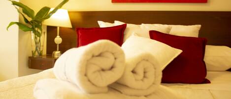 Una cama bien hecha con toallas blancas enrolladas y almohadas rojas, una mesita de noche con una lámpara, y un cuadro abstracto rojo en la pared.