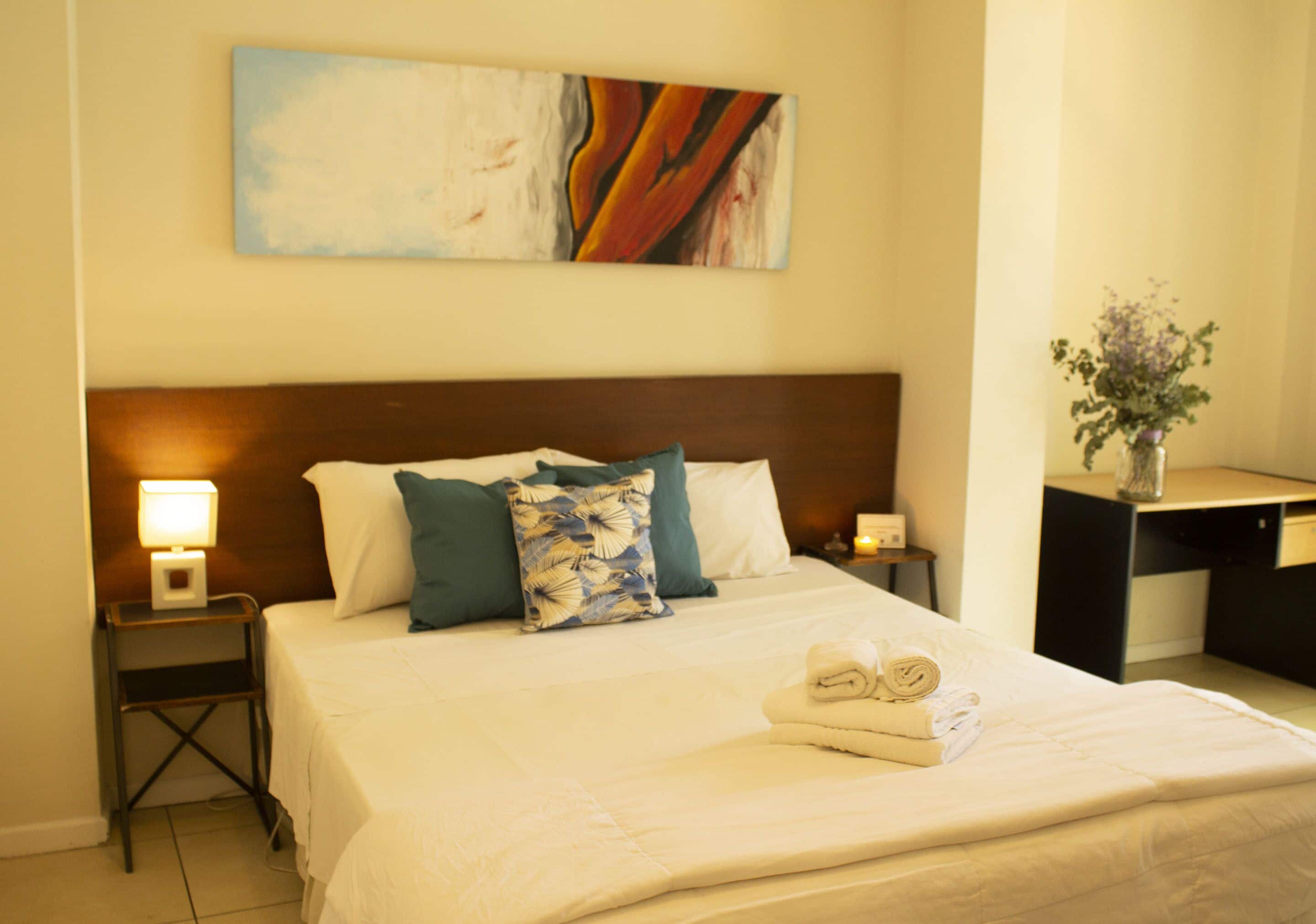 Una cama bien hecha con cabecera, almohadas y una toalla enrollada. Se ve una mesita de noche con una lámpara y una pequeña mesa con un florero.