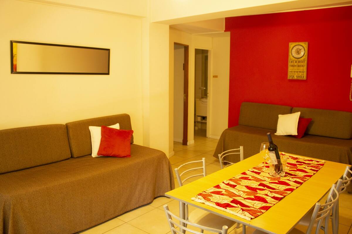 Una habitación con un sofá marrón, una pared roja y una mesa amarilla con mantel individual rojo y blanco.