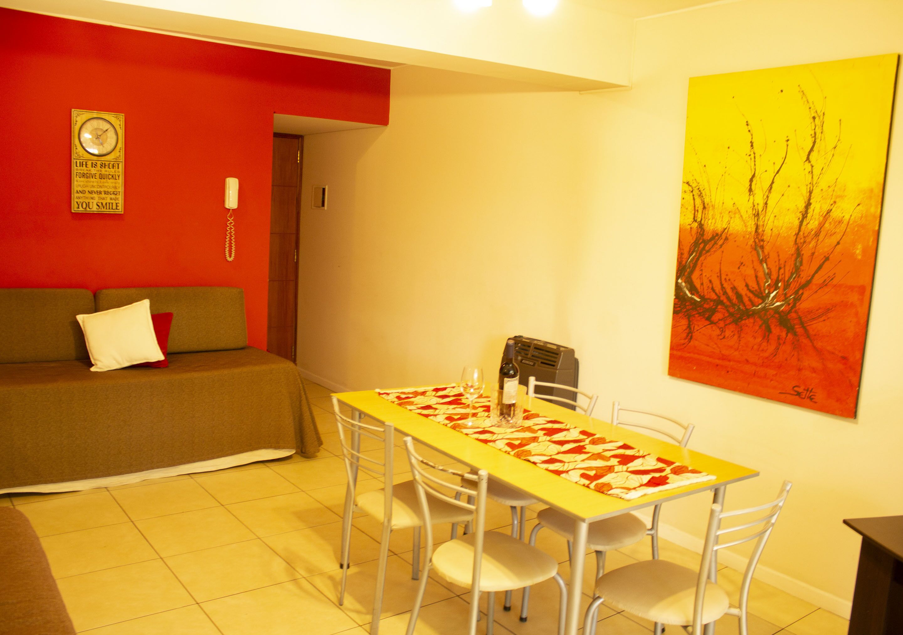 A dining area with a yellow table, white chairs, a red wall, and a painting.