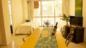 Habitación de hotel con una mesa de comedor, dos camas, un televisor y un amplio ventanal con cortinas.