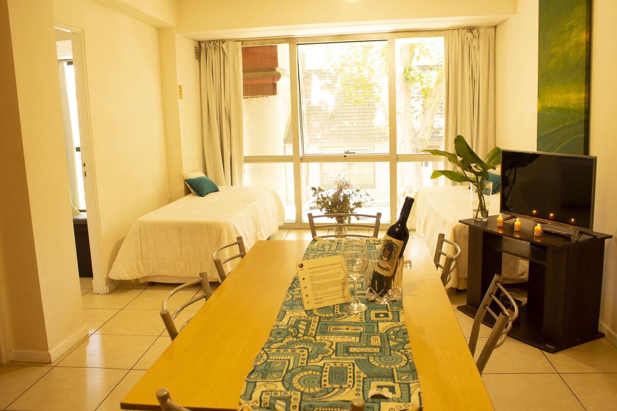 Habitación de hotel con una mesa de comedor, dos camas, un televisor y un amplio ventanal con cortinas.
