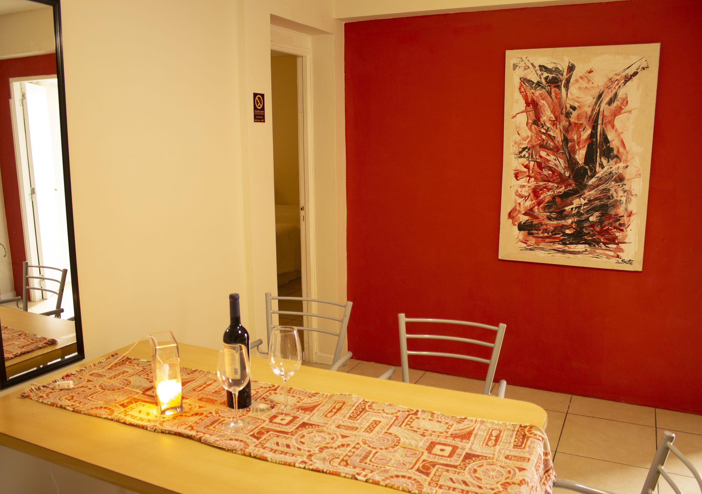 A dining area with a yellow table, wine glass, bottle, and candle. There is a red wall with a painting and a door in the background.