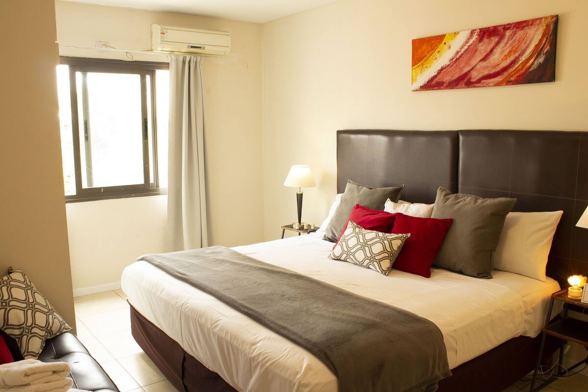 Habitación de hotel con una cama grande, una mesita de noche, una lámpara, una ventana con cortinas y un cuadro en la pared.