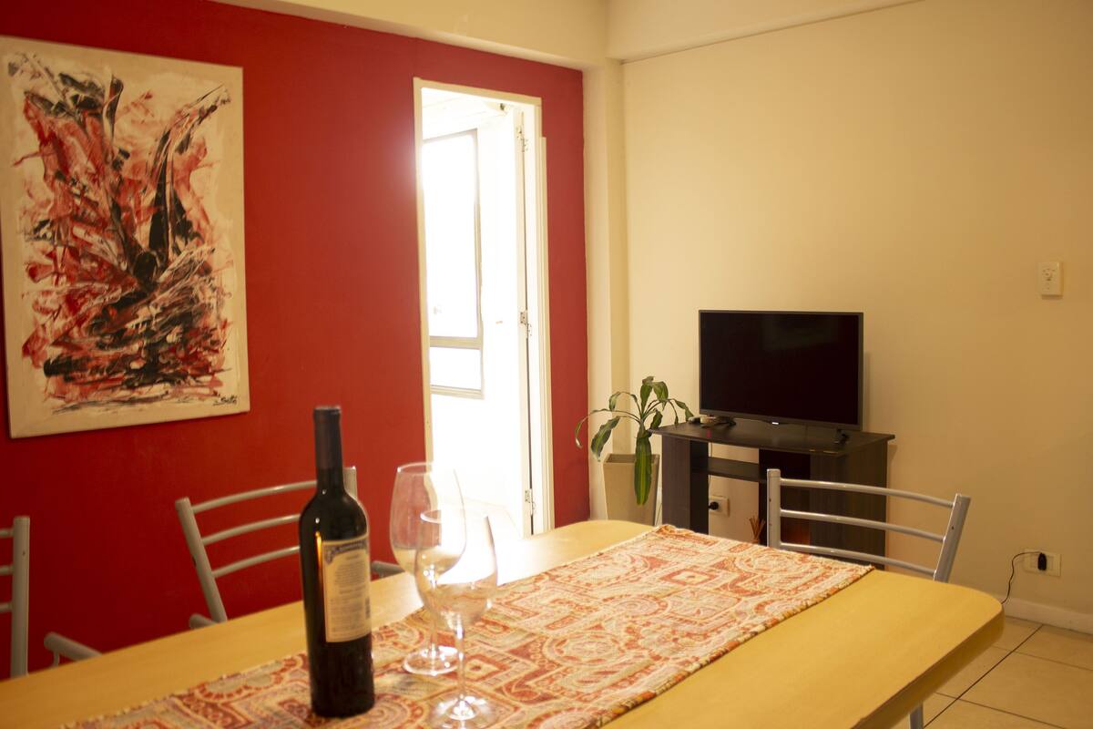 Un comedor con una mesa dispuesta para dos personas, una botella de vino y una copa. Hay un cuadro en la pared y un televisor sobre un mueble.