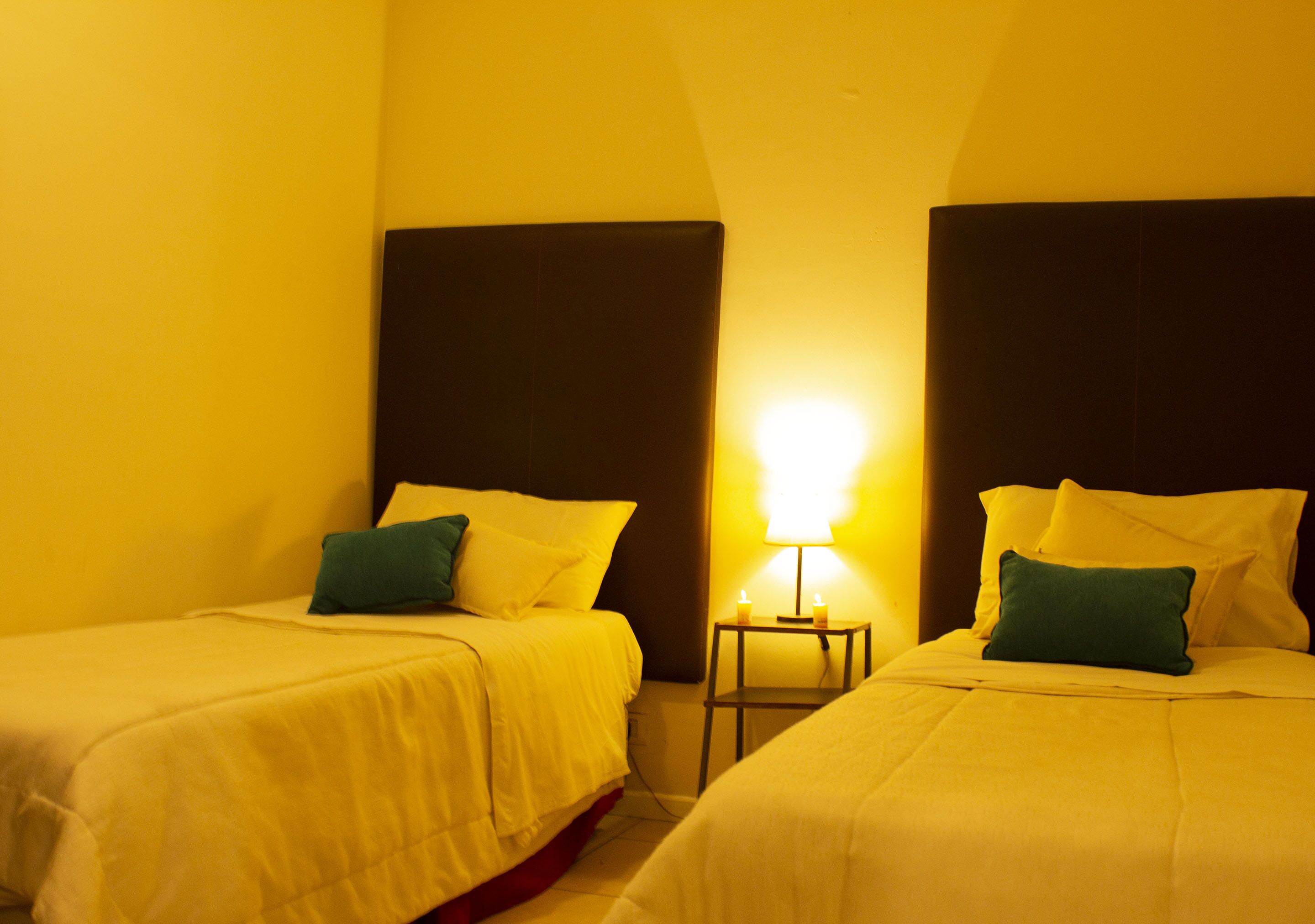 Two single beds with white bedding and green pillows, a small bedside table with a lamp, and a yellow wall.