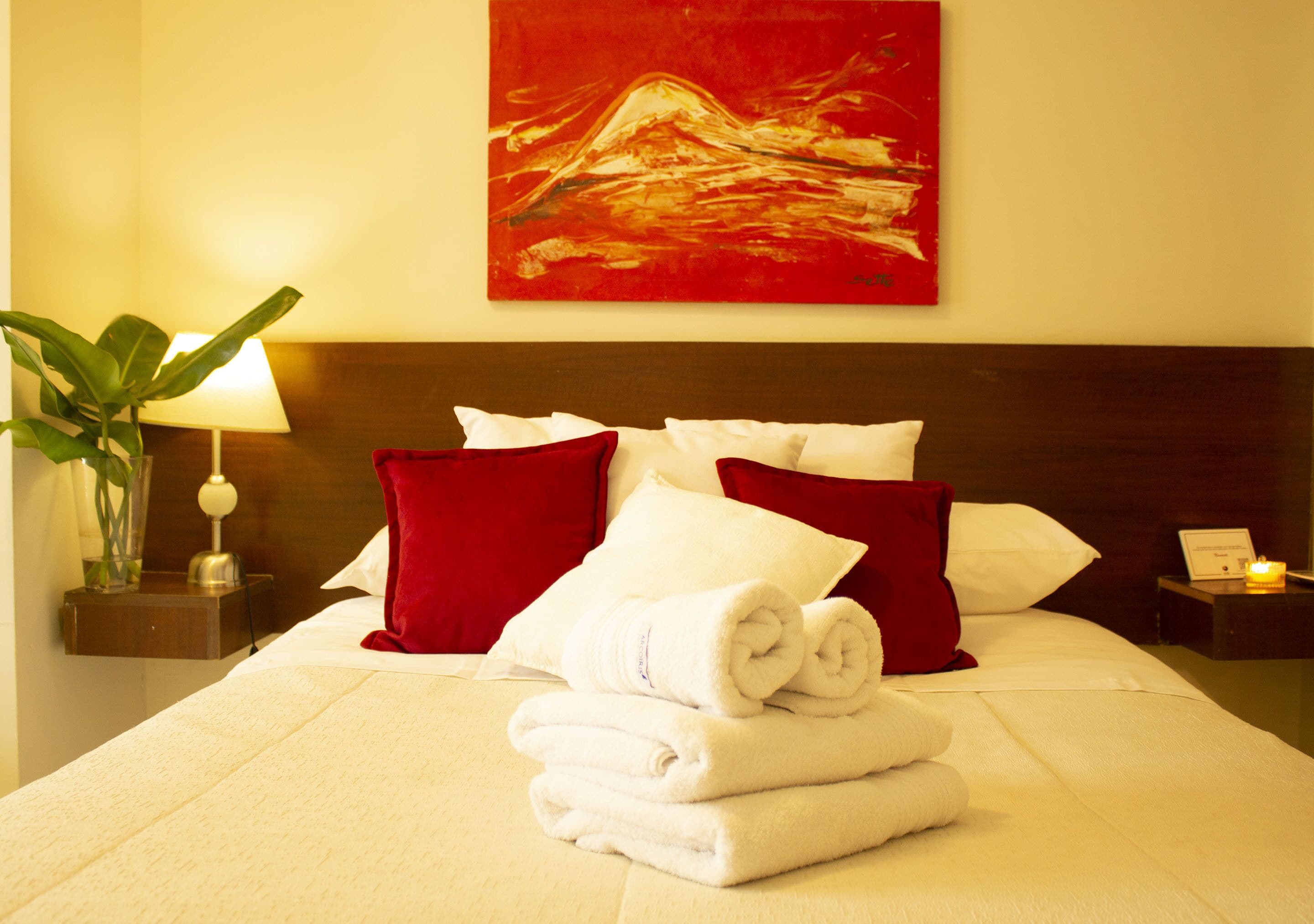 Une chambre d’hôtel avec un lit soigneusement fait, des oreillers rouges et blancs, une œuvre d’art rouge accrochée au mur et une lampe de chevet.