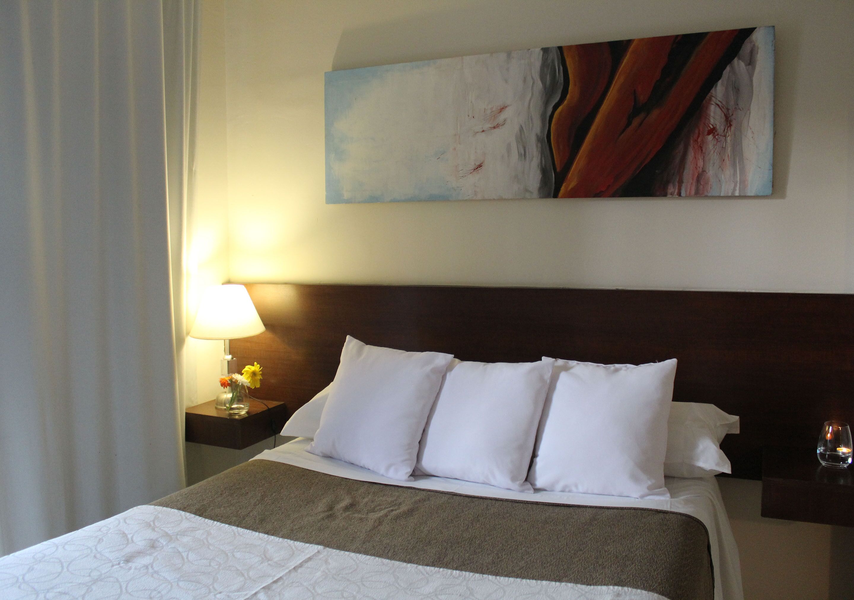 A hotel room with a neatly made bed, a painting on the wall, a nightstand with a lamp and a vase of flowers.