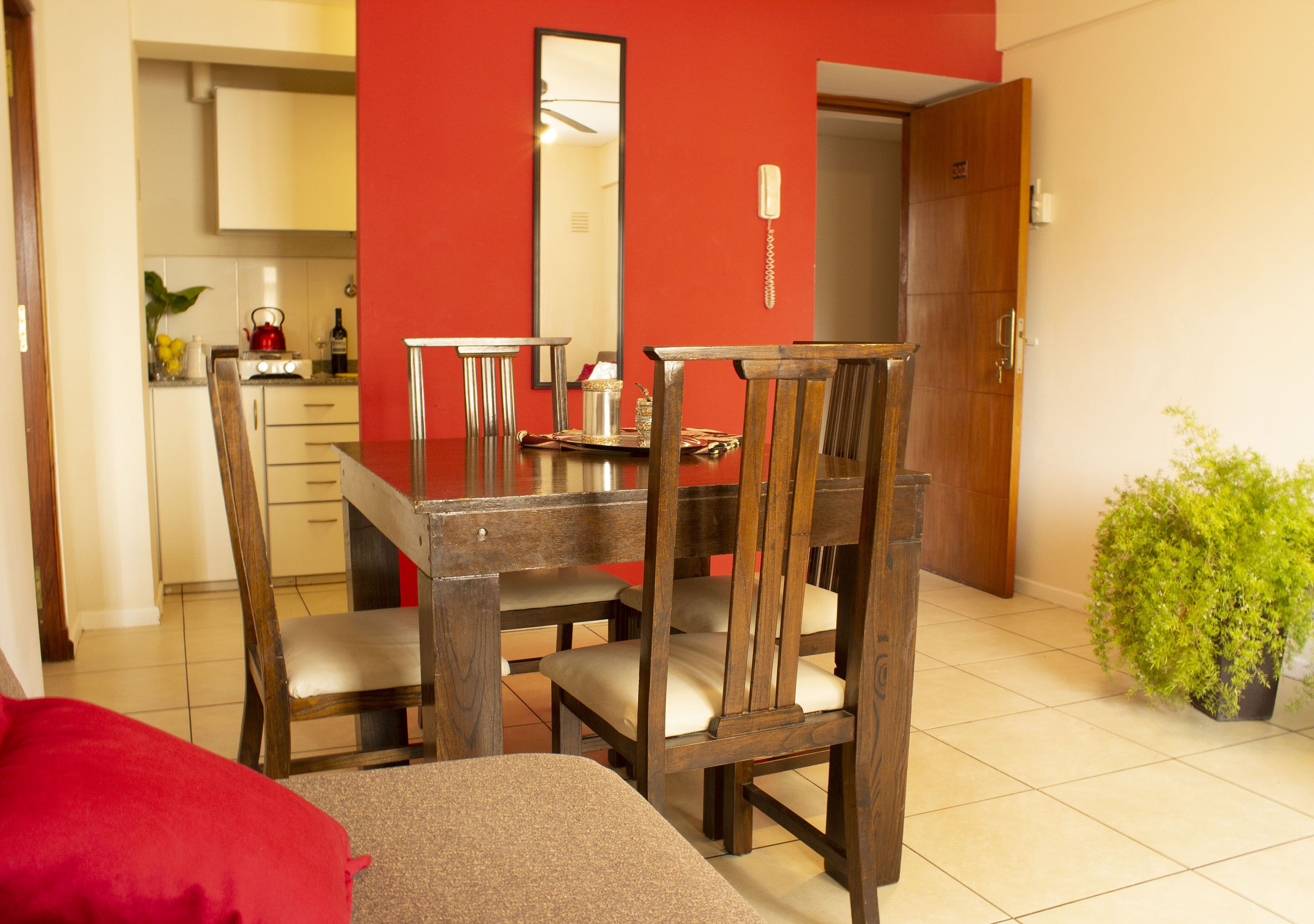 A dining area with a wooden table and chairs, a red accent wall, a kitchen in the background, and a potted plant.