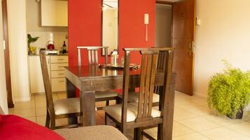 A dining area with a wooden table and chairs, a red accent wall, a kitchen in the background, and a potted plant.