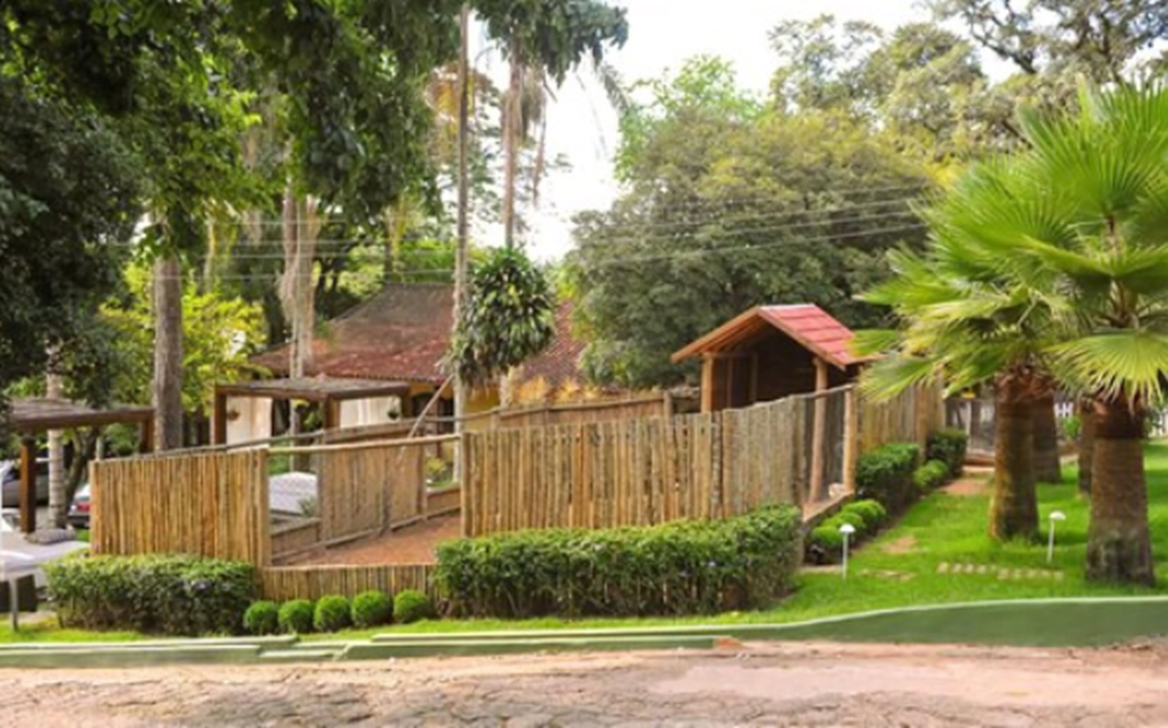 Uma cerca de madeira rústica, uma pequena construção de madeira e um gramado bem cuidado com uma palmeira.