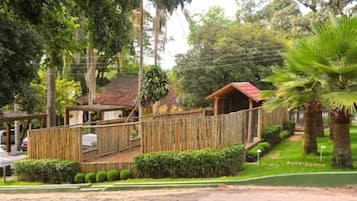 Uma cerca de madeira rústica, uma pequena construção de madeira e um gramado bem cuidado com uma palmeira.