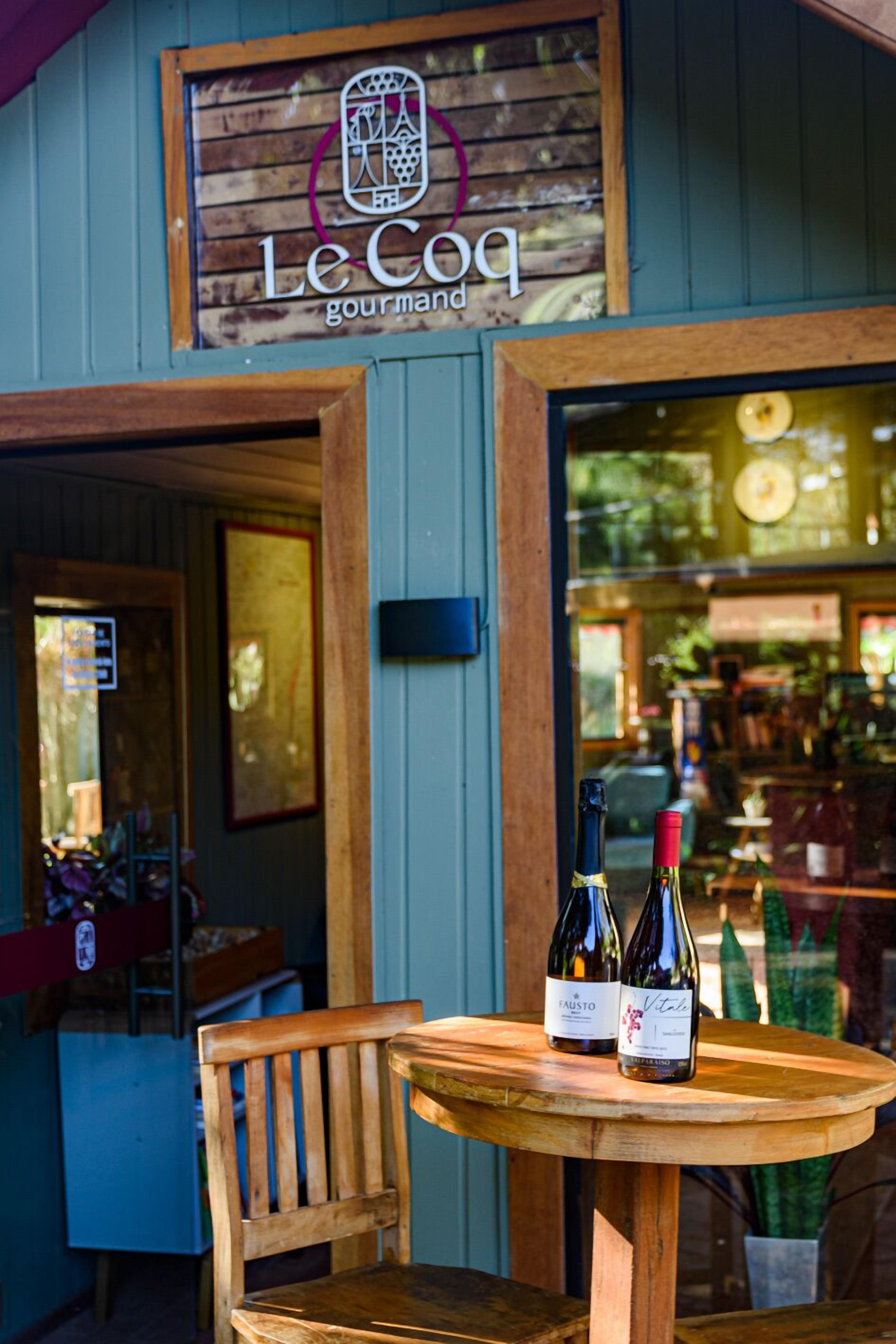 Une table en bois avec deux bouteilles de vin devant un bâtiment sur lequel on peut lire l’inscription « Le Coq gourmet ».