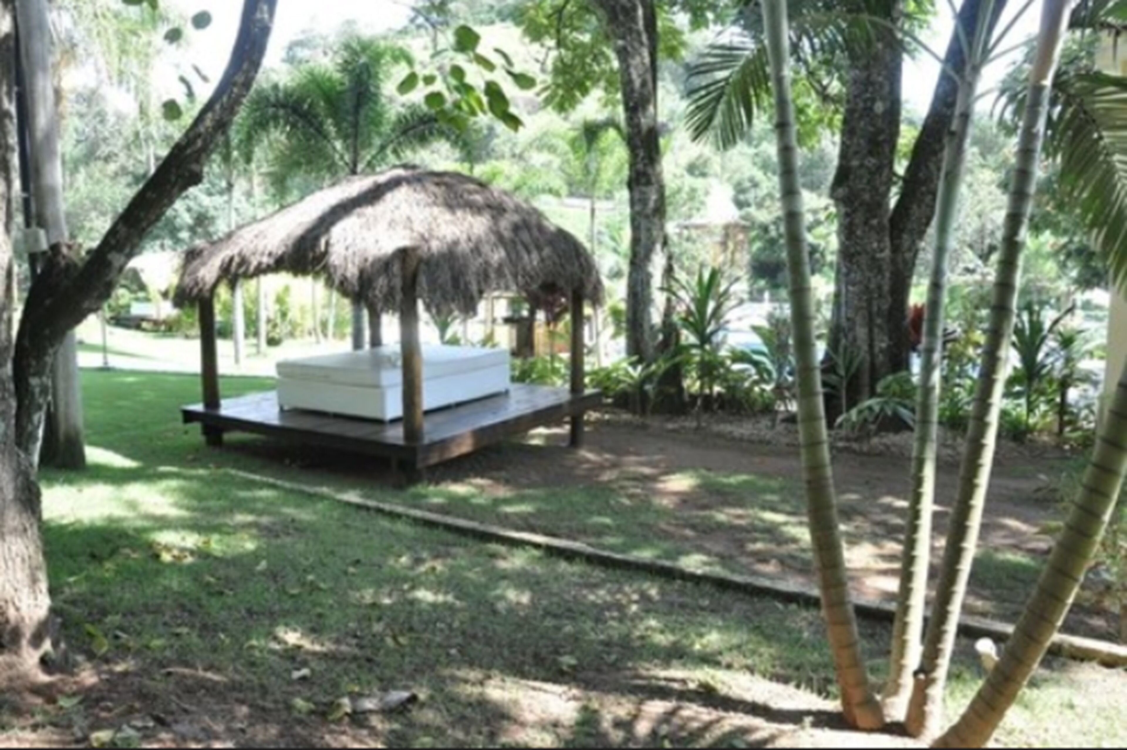 A thatched-roof outdoor seating area with a cushioned bench, surrounded by lush greenery and trees.