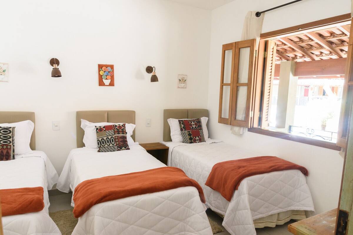 Ein Hotelzimmer mit drei Einzelbetten, jedes mit weißer Bettdecke und orangefarbener Tagesdecke. Wandlampen und eine Uhr sind an der Wand angebracht.