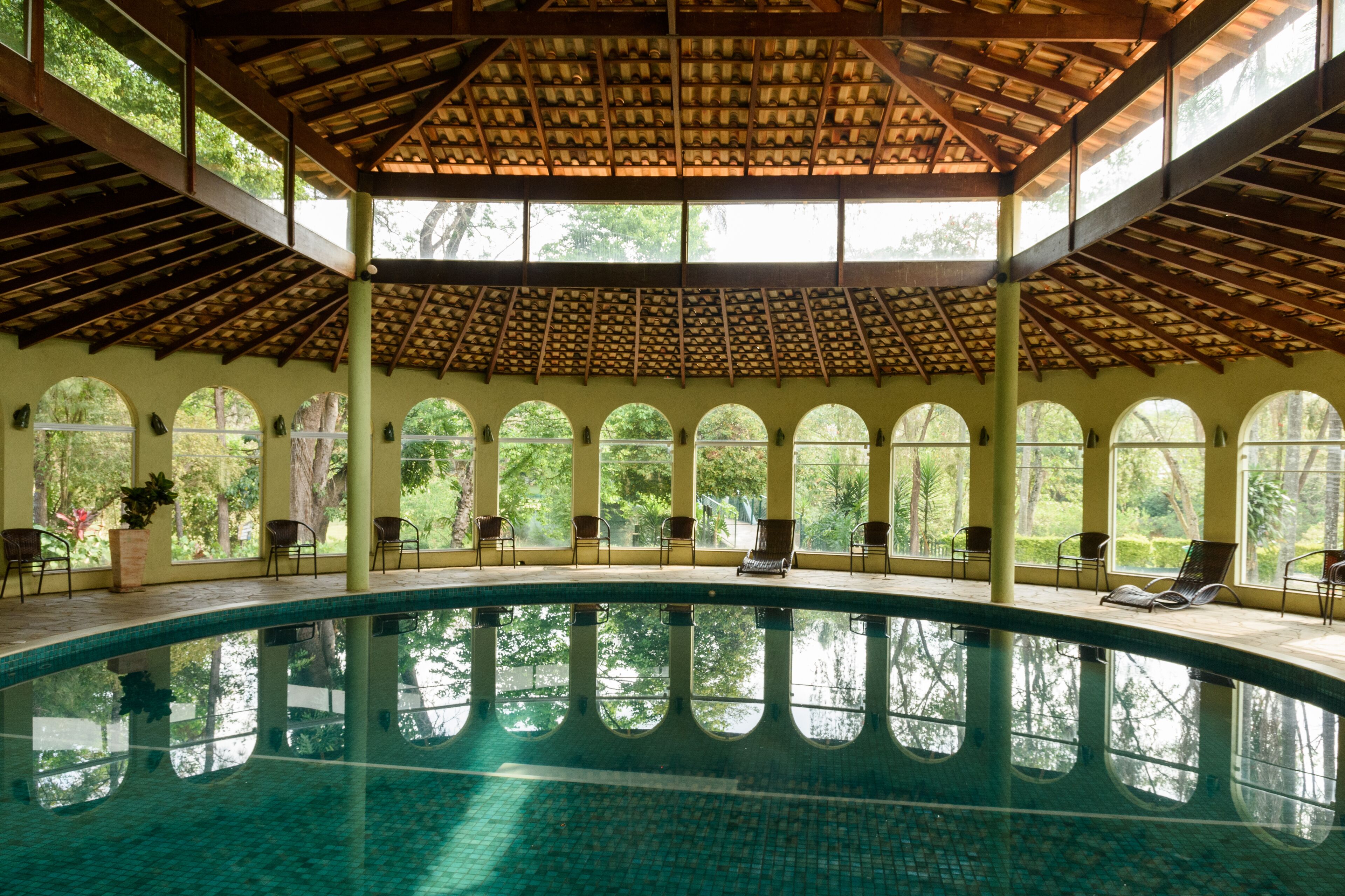 An indoor swimming pool with a tiled floor, surrounded by arched windows and chairs.