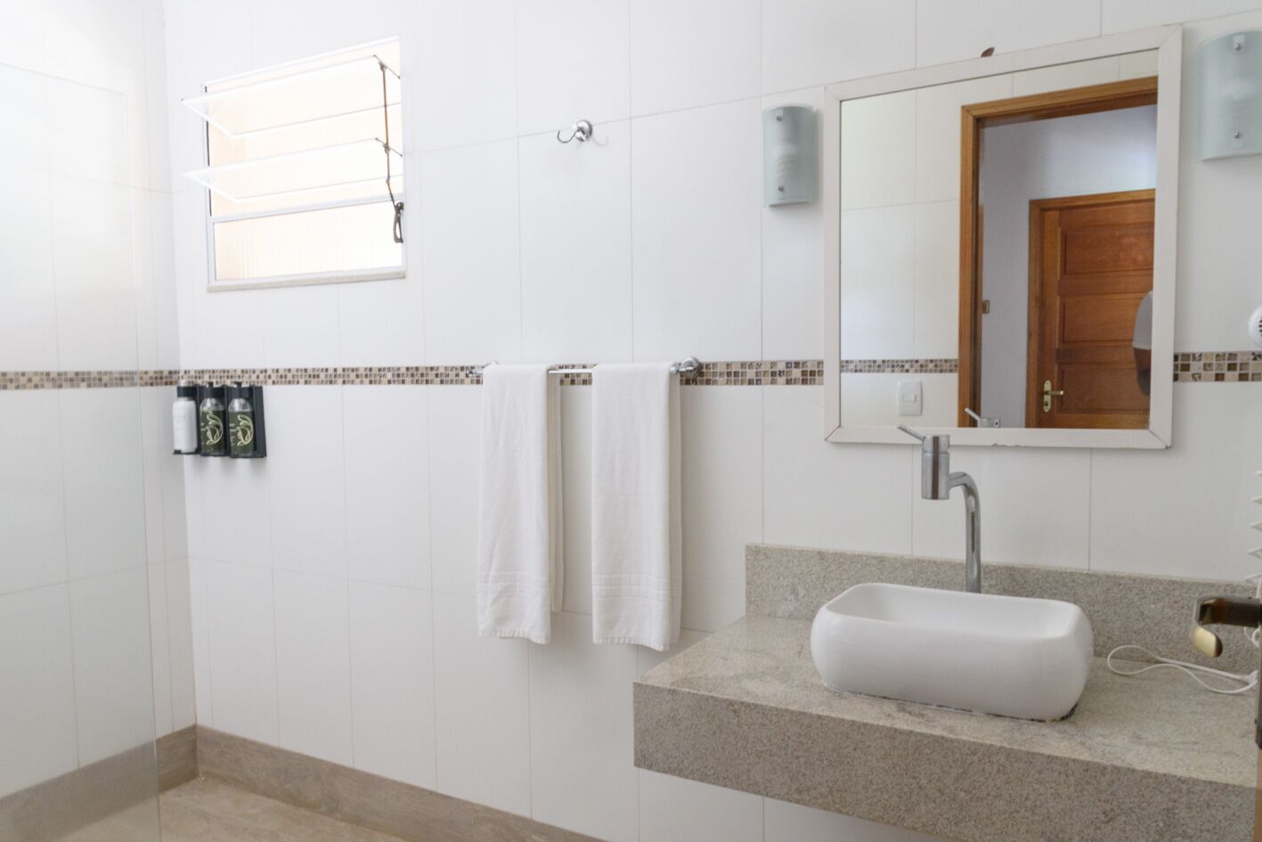 A bathroom with white tiles, a sink, a mirror, and a window with blinds.