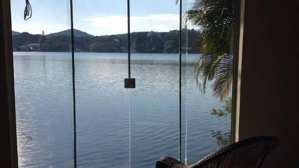 A room with a large sliding glass door leading to a lake view, a wicker chair, and a hanging light fixture. - Koze Hostel (Florianópolis)