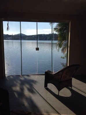 A room with a large sliding glass door leading to a lake view, a wicker chair, and a hanging light fixture. - Koze Hostel (Florianópolis)