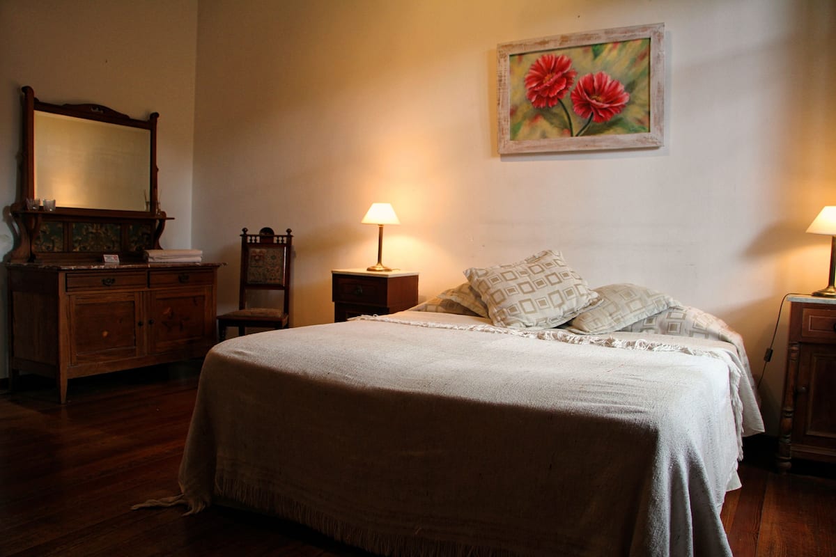 A bedroom with a large bed, two bedside tables with lamps, a dresser, a chair, and a painting on the wall.