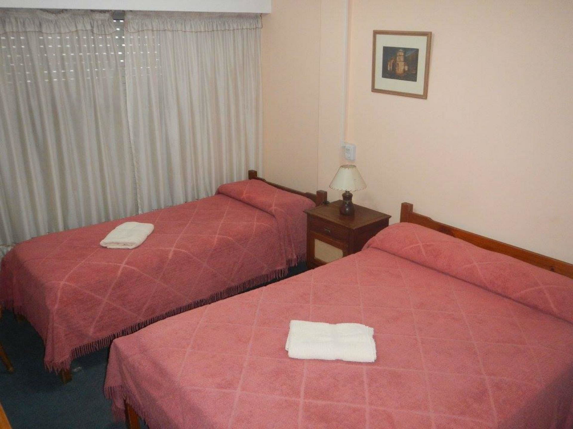 Una habitación de hotel con dos camas individuales, cada una con una colcha rosa y una toalla blanca doblada encima. Hay una mesita de noche con una lámpara entre las camas.
