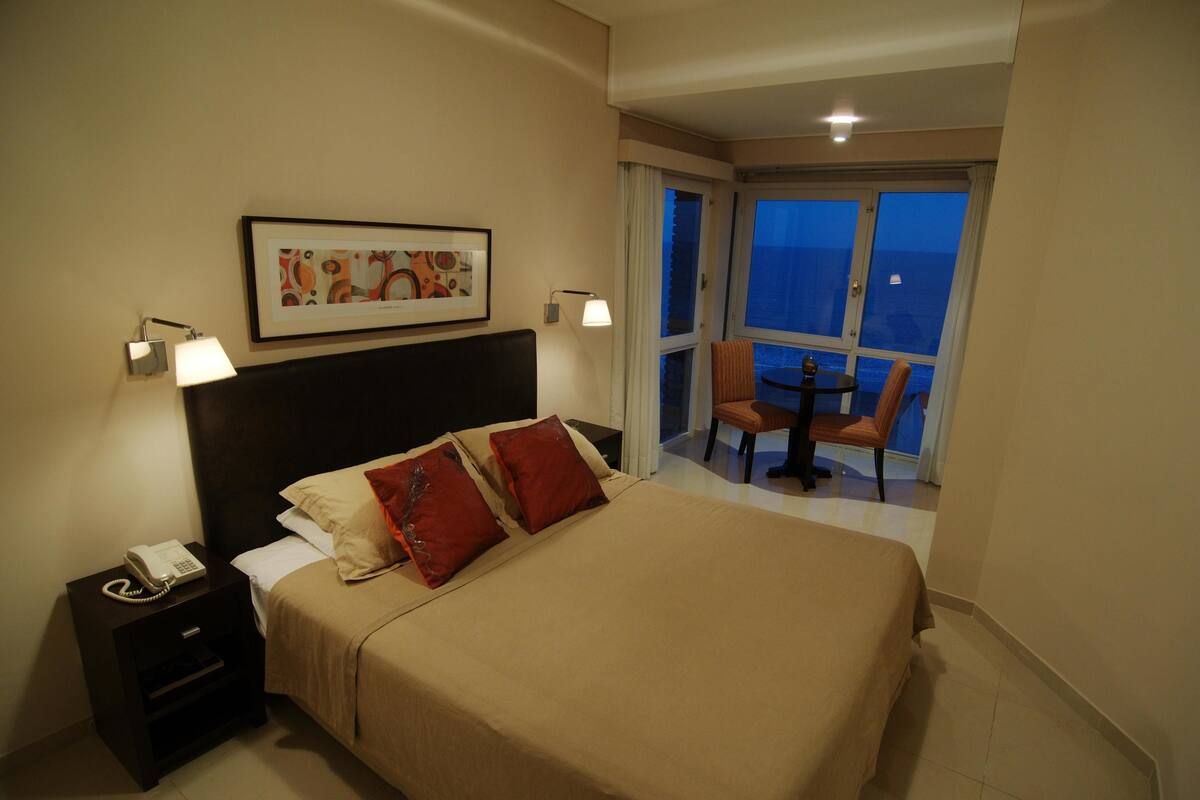 A hotel room with a bed, a nightstand with a phone, a framed picture on the wall, and a balcony with a table and chairs.