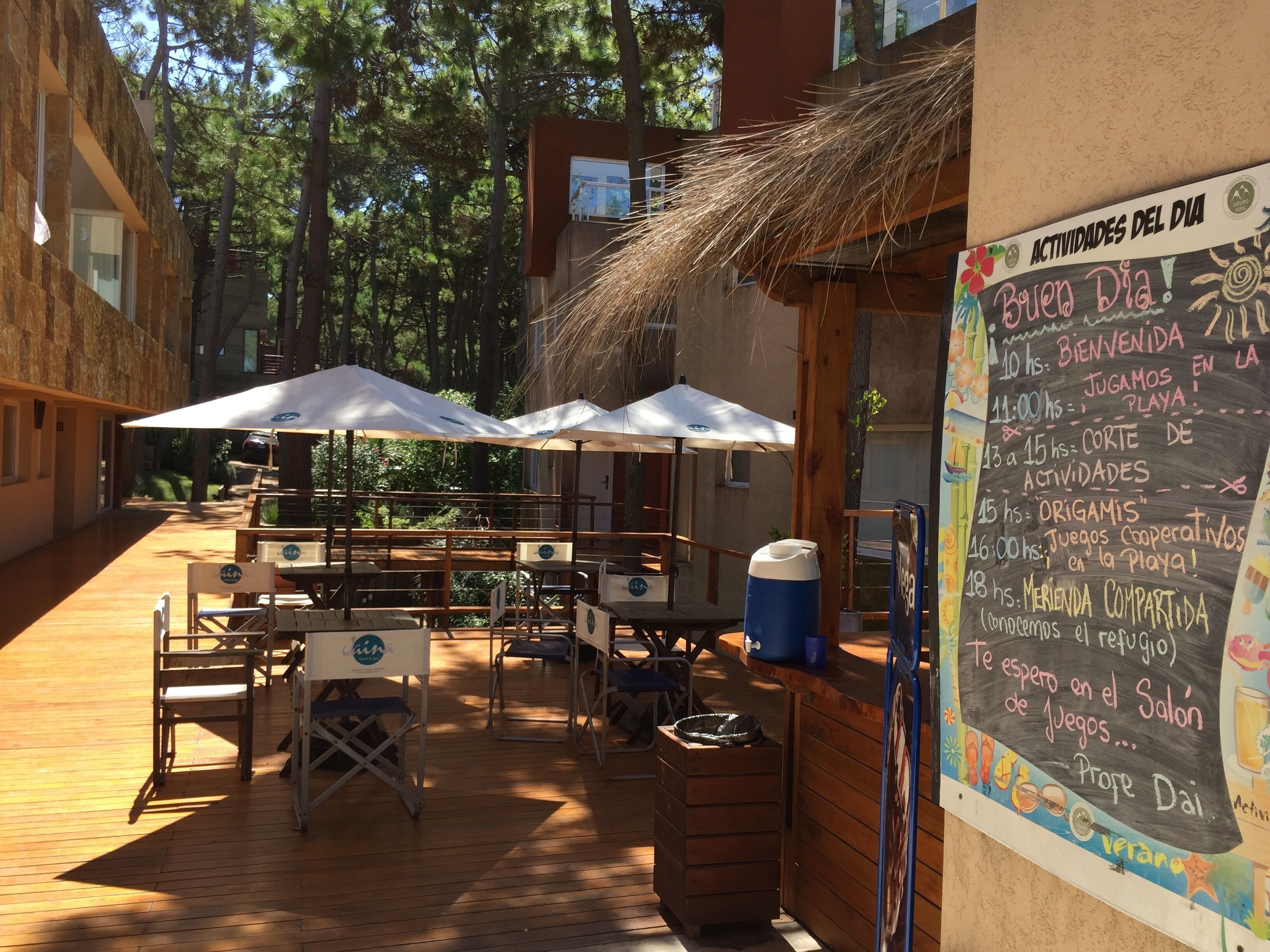 An outdoor seating area with tables and umbrellas, a chalkboard with daily activities, and a wooden deck.