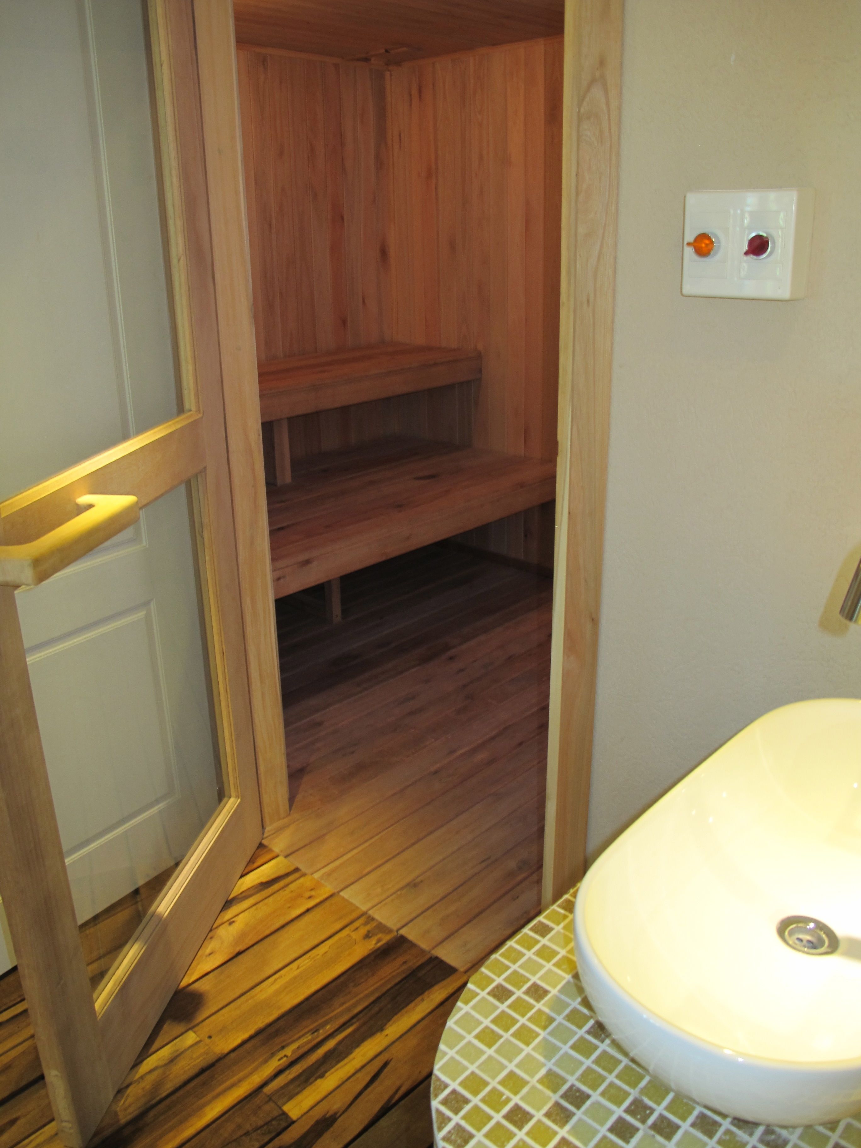 A wooden sauna with a bench, a white sink with a tiled basin, and a wall-mounted light switch.