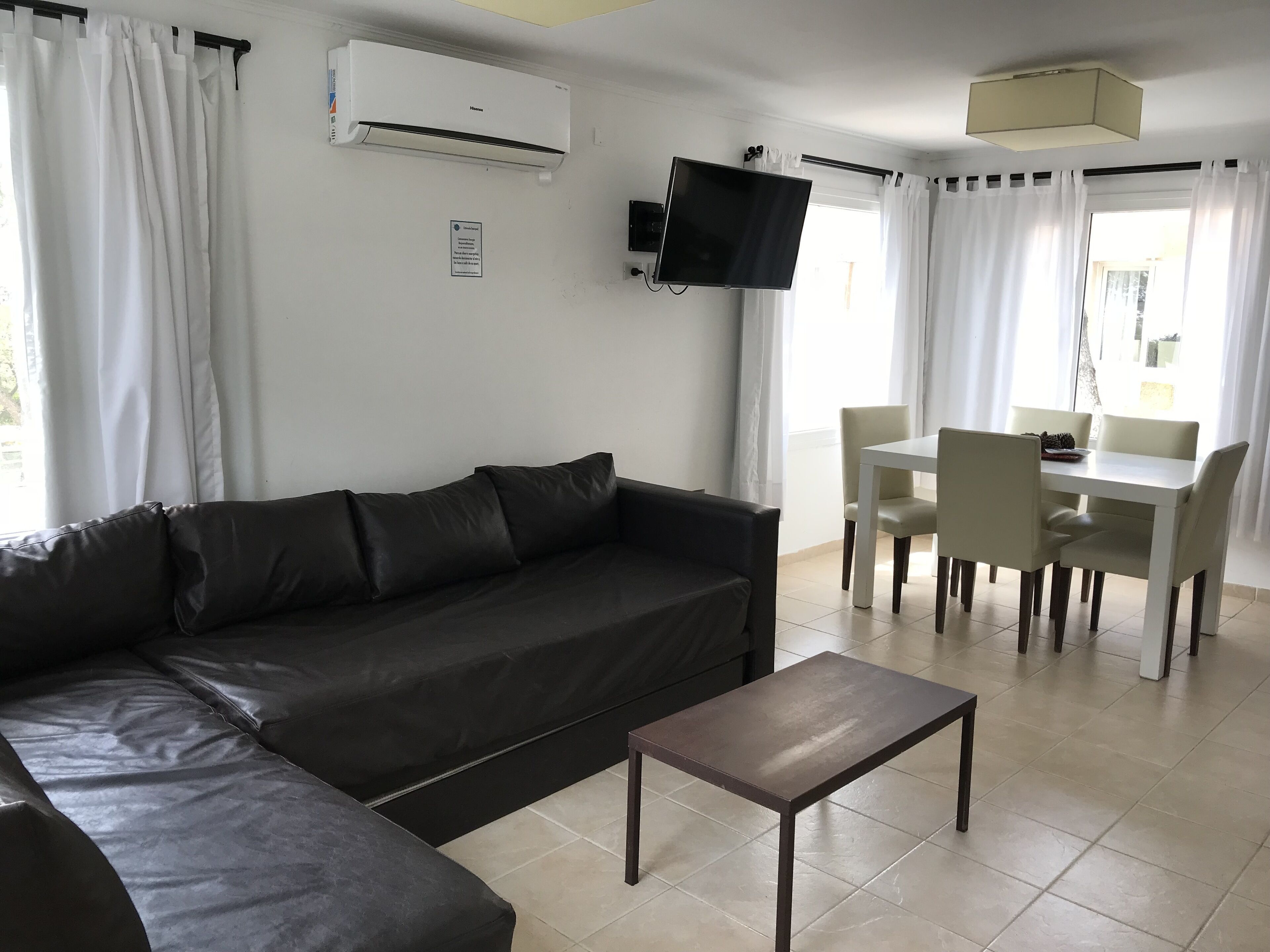 A living room with a black leather sofa, a small wooden table, a TV mounted on the wall, and a dining area with a white table and chairs.