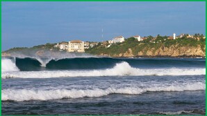 Paisaje costero con olas rompiendo en la orilla, edificios en un acantilado y un faro al fondo.