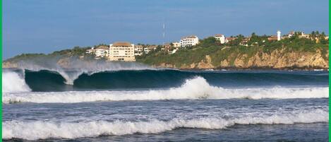 Paisaje costero con olas rompiendo en la orilla, edificios en un acantilado y un faro al fondo.