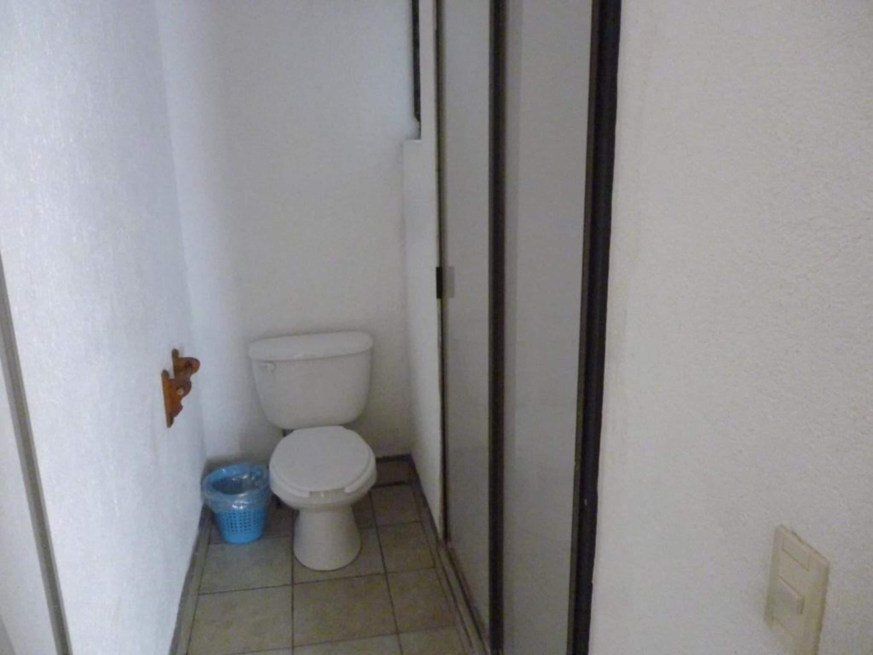 Un baño pequeño y estrecho con un inodoro blanco y un cubo de basura azul.