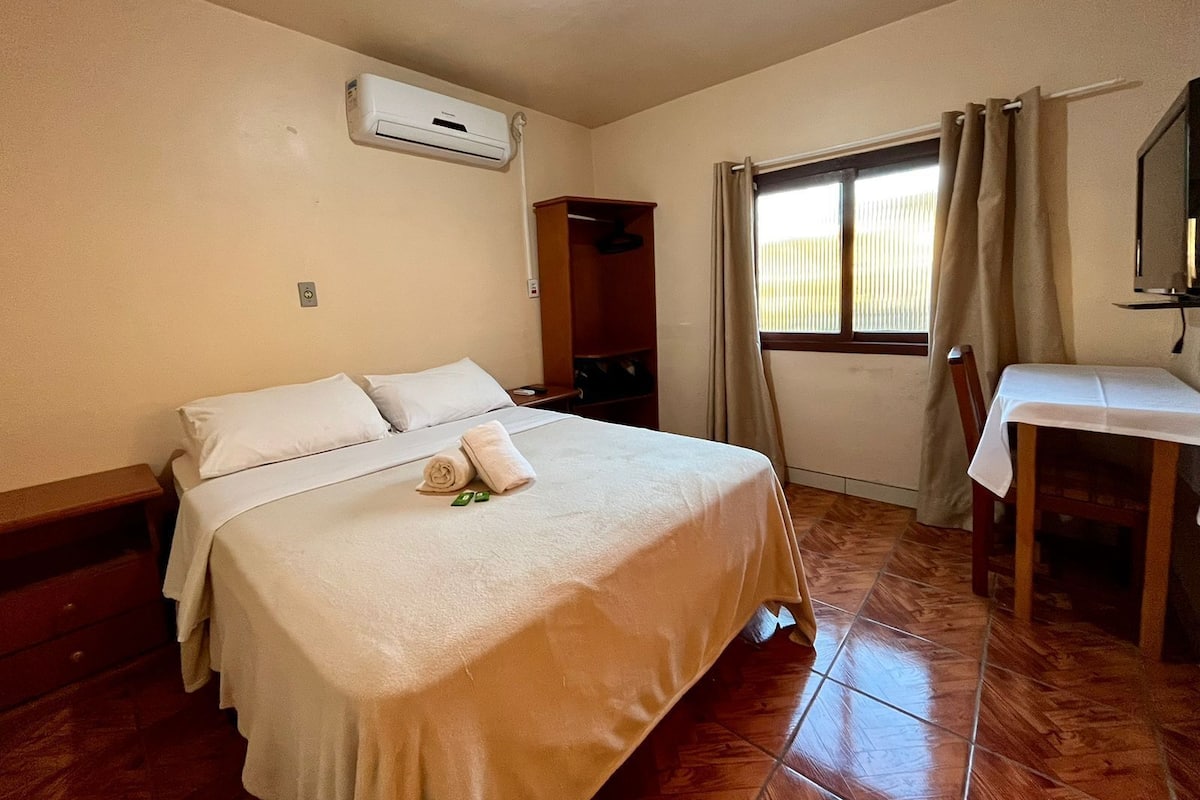 Quarto de hotel com cama, escrivaninha, ar condicionado e janela com cortinas.