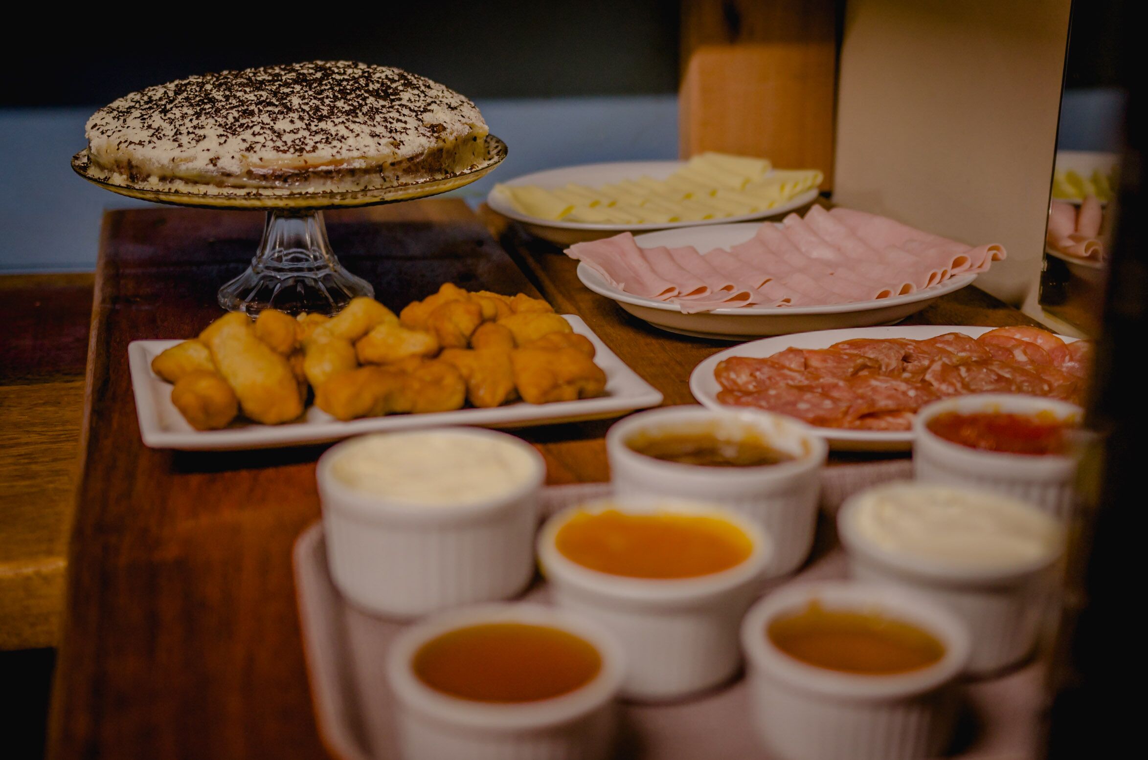 Uma mesa com diversos alimentos, incluindo torta, queijo, presunto e molhos para acompanhar.