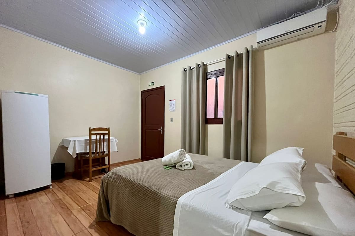 Quarto de hotel com cama, uma pequena mesa com cadeira, um refrigerador e uma janela com cortinas.