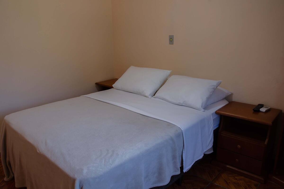 Uma cama de solteiro com um cobre-leito branco e duas almofadas, uma mesa de cabeceira de madeira com um telefone e uma tomada.