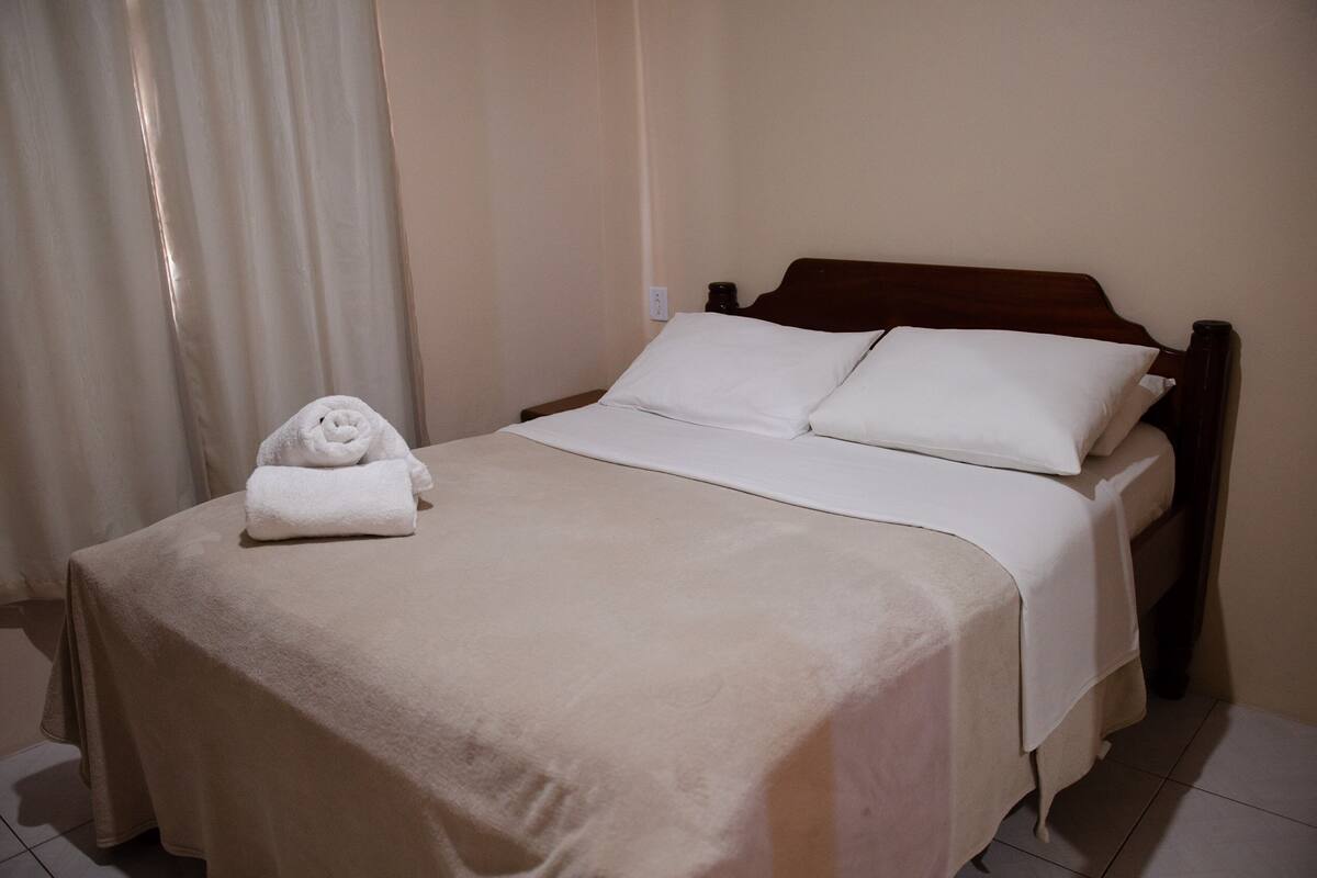 Cama bem arrumada com colcha bege e almofadas, uma toalha enrolada sobre a cama e cabeceira de madeira.