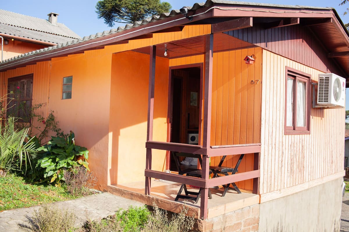 Uma casa de madeira com varanda, uma construção pequena anexa e uma janela com unidade de ar condicionado.