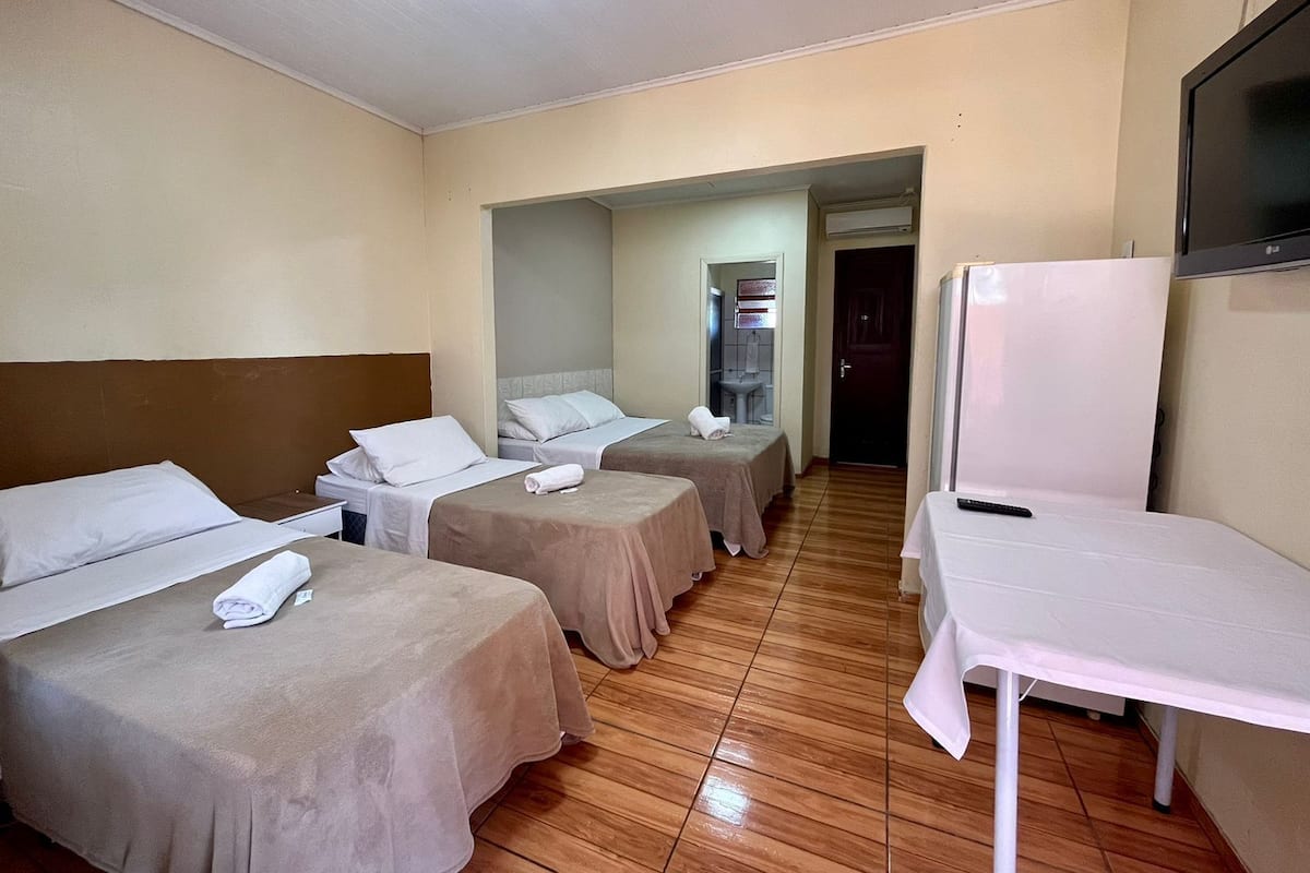 Quarto de hotel com três camas simples, uma mesa pequena, televisão e banheiro visível por um arco.