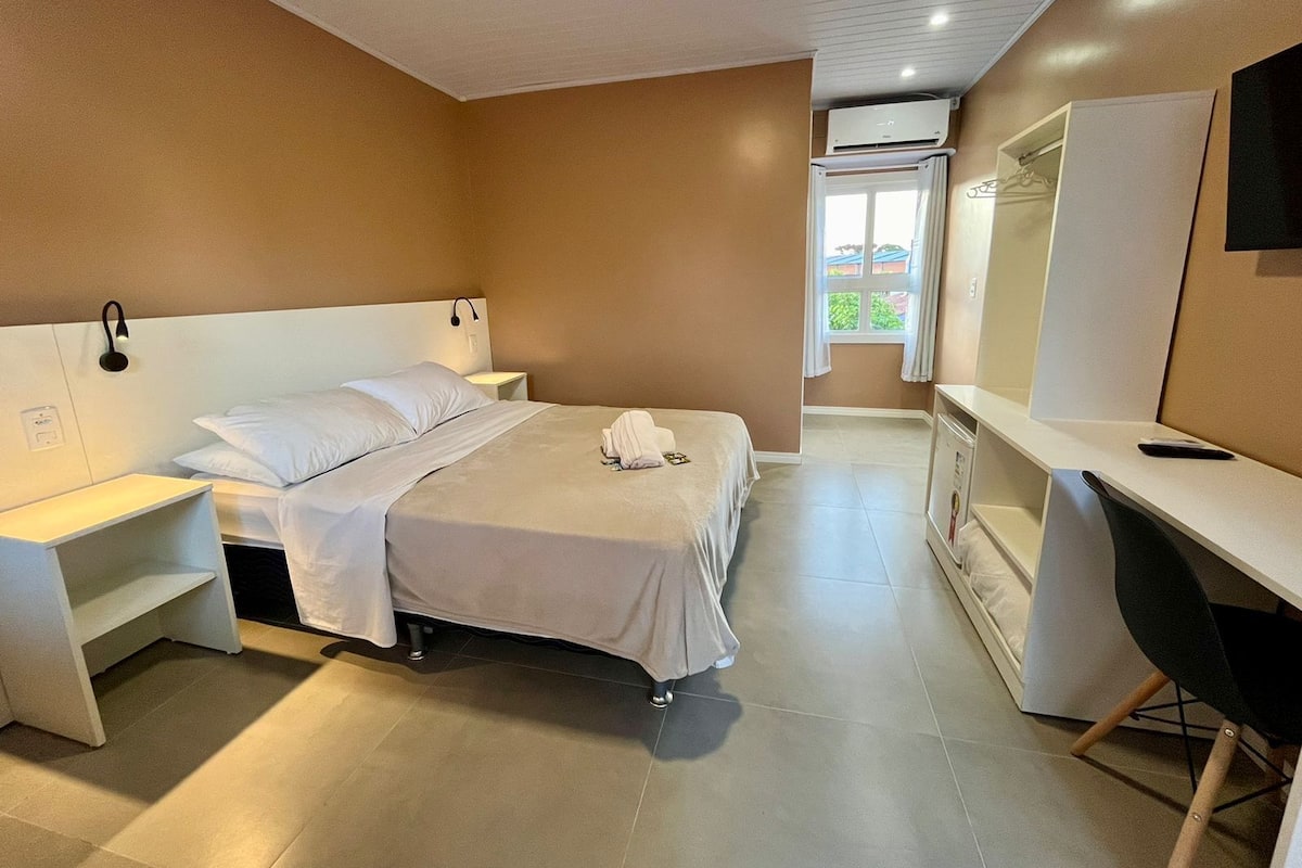 Quarto de hotel com cama, mesas de cabeceira, escrivaninha, cadeira, televisão e ar condicionado.