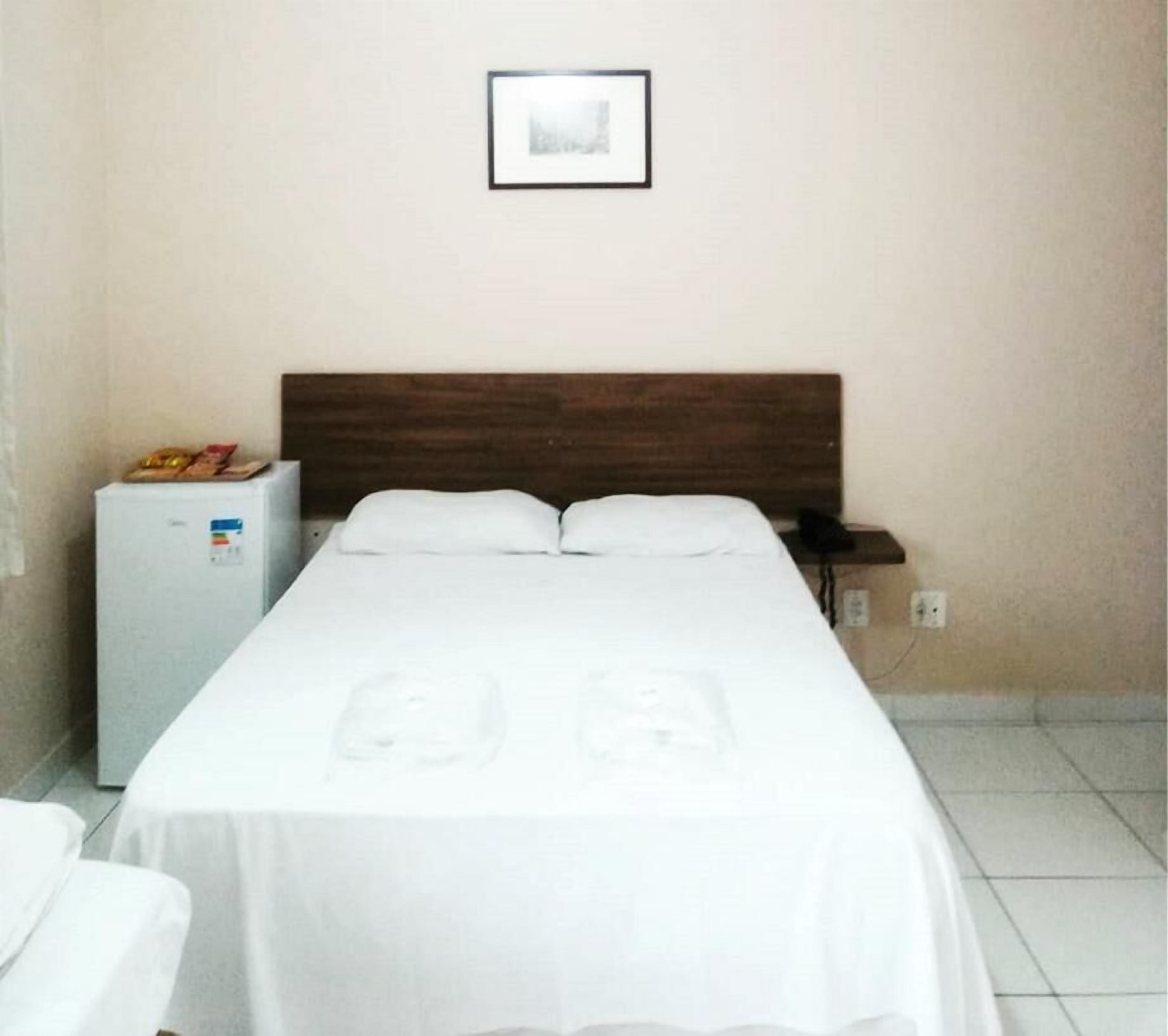 Habitación de hotel con cama, mesita de noche, refrigerador pequeño y un cuadro en la pared.