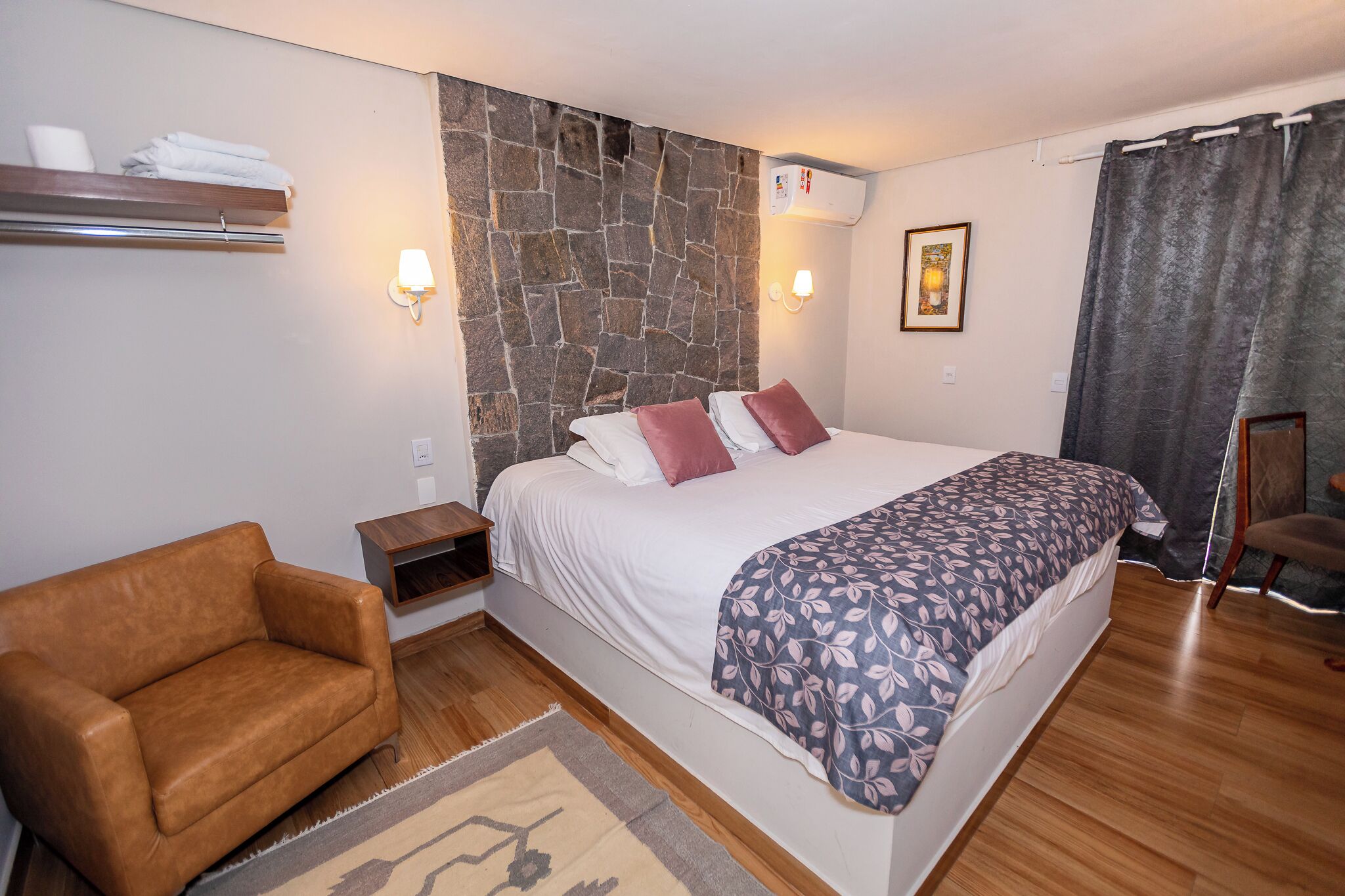 A bedroom with a large bed, a brown leather chair, a stone accent wall, a bedside table, and a wall-mounted shelf with towels.